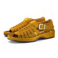 A pair of open-toe sandals in mustard yellow suede, featuring a braided front strap, a side buckle strap, and a chunky sole, shown on a white background.
