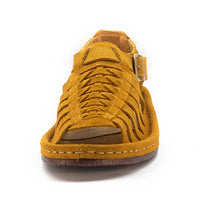 A single strappy sandal in mustard yellow suede, featuring a buckle strap, braided front straps, and a wedge sole, shown against a plain white background.